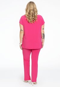 Fuchsia todeltøj med en løs top og lige bukser, lavet af glat stof. Bemærkelsesværdige tatoveringer på armen og hvide accessories.