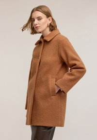 Brązowy wełniany płaszcz z usztywnionym kołnierzem, bocznymi kieszeniami i guzikami, z teksturowanym wykończeniem i średniej długości, luźną sylwetką.