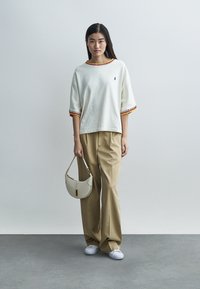 Bela teksturirana bluza z rdečimi in rumenimi poudarki na rokavih, kombinirana z bež širokimi hlačami, drži kremno okroglo torbico in nosi bele čevlje.