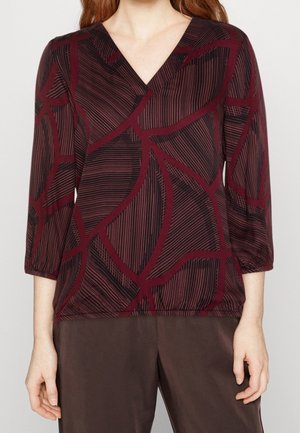 Vrouw draagt een bordeauxrode V-hals blouse met abstracte donkerrode en zwarte gebogen lijnpatronen en driekwart mouwen, gecombineerd met donkere broek.