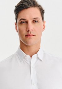 Młody mężczyzna z jasnobrązowymi włosami i zielonymi oczami, ubrany w białą koszulę zapinaną na guziki, patrzy prosto przed siebie na tle jednolitego białego tła.