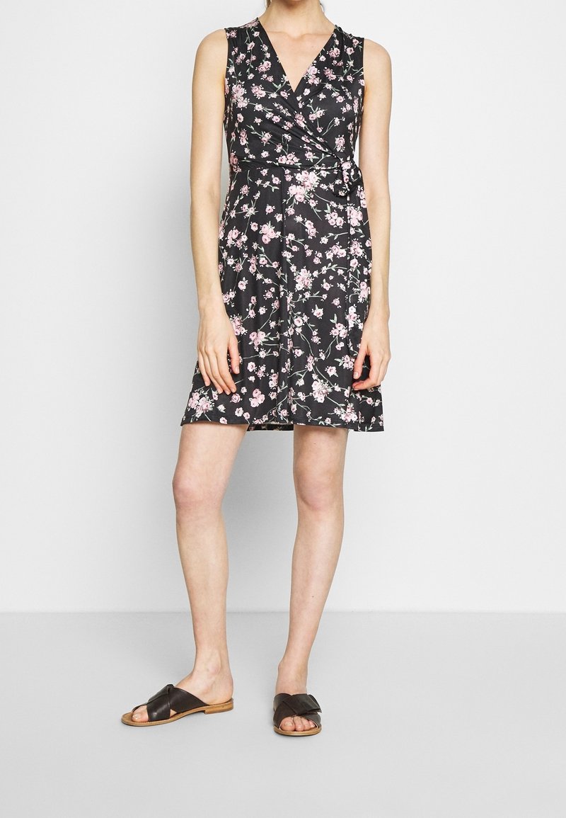 Femme portant une robe portefeuille noire sans manches avec un imprimé floral rose et des sandales plates noires à bouts ouverts, debout devant un fond blanc uni.