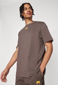 T-shirt marron à manches courtes avec un col rond, arborant un logo jaune sur la poitrine. Fait en tissu doux, avec une coupe décontractée et un design casual.