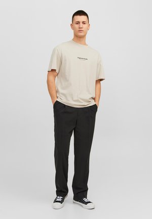 Beige cotton t-shirt with short sleeves and a printed logo, paired with black trousers and black sneakers, standing against a neutral background.