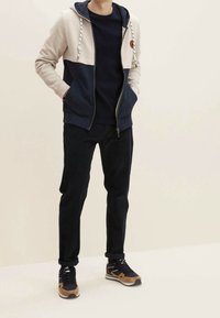 Persona che indossa una felpa con zip beige e blu navy, pantaloni neri e scarpe da ginnastica nere e marroni, in piedi contro uno sfondo chiaro e semplice.
