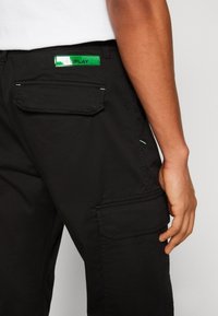 Pantaloni cargo neri con due tasche laterali, un'etichetta del marchio verde sul retro e dettagli di cucitura a contrasto. Il materiale sembra resistente e liscio.