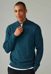 Türkisfarbener Zip-Pullover aus strukturiertem Stoff, mit langen Ärmeln und lockerem Schnitt, über einem weißen T-Shirt getragen.