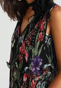 Abito plissettato floreale con sfondo nero, fiori vivaci in rosso, viola e verde, con orlo arricciato e un accento decorativo a forma di fiore.