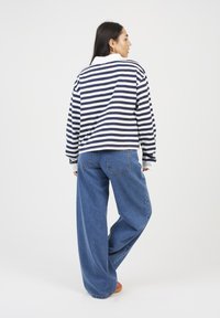 Woman with long dark hair stands facing sideways, wearing a white and navy striped long-sleeve shirt, wide-leg blue jeans, and orange shoes.