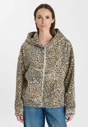 Vrouw draagt een hoodie met volledige rits en luipaardprint met voorzakken en blauwe jeans, staand tegen een witte effen achtergrond.