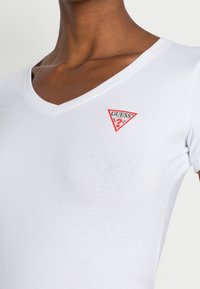 Biała bawełniana koszulka z dekoltem w serek. Posiada małe czerwone trójkątne logo na lewej stronie klatki piersiowej, gładką fakturę, krótkie rękawy.