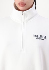 Vit zip-up sweatshirt med hög krage, med marinblå "Hollister California" brodyr på bröstet. Mjuk texturerad tyg.