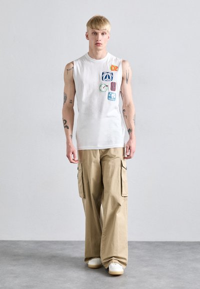 Tricou alb fără mâneci cu insigne decorative, asortat cu pantaloni cargo bej lejeri, prevăzuți cu buzunare laterale, și adidași albi.