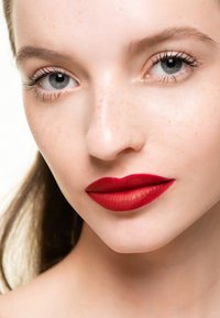 Primer plano de un rostro con piel clara y pecas, con ojos azules y labios llenos cubiertos de lápiz labial rojo mate. Se puede observar una textura de piel suave.