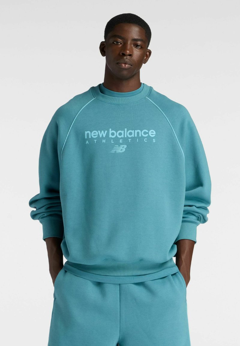 Giovane uomo con le mani in tasca che indossa una felpa teal New Balance Athletics e pantaloni abbinati, su sfondo bianco semplice.
