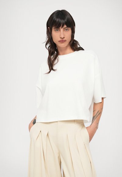 Λευκό κοντομάνικο crop top με στρογγυλή λαιμόκοψη, συνδυασμένο με μπεζ πλισέ παντελόνι. Λείο ύφασμα, μινιμαλιστικός σχεδιασμός.