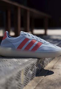 Zapatilla deportiva azul clara con tres rayas rojas descansando sobre una superficie metálica texturizada al aire libre bajo una luz solar suave.