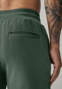 Green textured joggers with a ribbed waistband and a small, flat back pocket. The fabric appears breathable and durable.