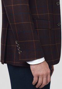 Brązowa marynarka w kratę z niebiesko-pomarańczowym wzorem, fakturowana tkanina, trzy guziki przy mankiecie oraz widoczny mankiet białej koszuli poniżej.