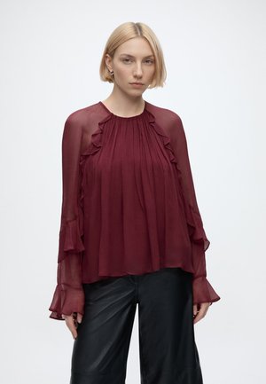Vrouw met kort blond haar draagt een bordeauxkleurige doorzichtige blouse met ruches en zwarte leren broek, staand tegen een effen lichte achtergrond.