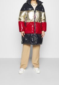 Puffjacka med glänsande svarta, guldiga och röda paneler; har en hög krage, dragkedjestängning och sidofickor; tillverkad av glänsande material.