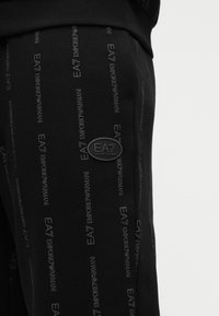 Pantalones jogger negros con un patrón repetitivo del logo "EA7 EMPORIO ARMANI", con una textura suave y un acento de logo circular en el lateral.