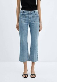 Jeans de mezclilla de corte acampanado en azul claro con dobladillo sin rematar y bolsillos delanteros, combinados con sandalias negras. La tela parece suave con una ligera textura.
