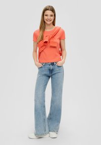 Koraalkleurig t-shirt met korte mouwen en ruches, gecombineerd met lichtblauwe flared jeans en witte sneakers. Soepele textuur en casual pasvorm.