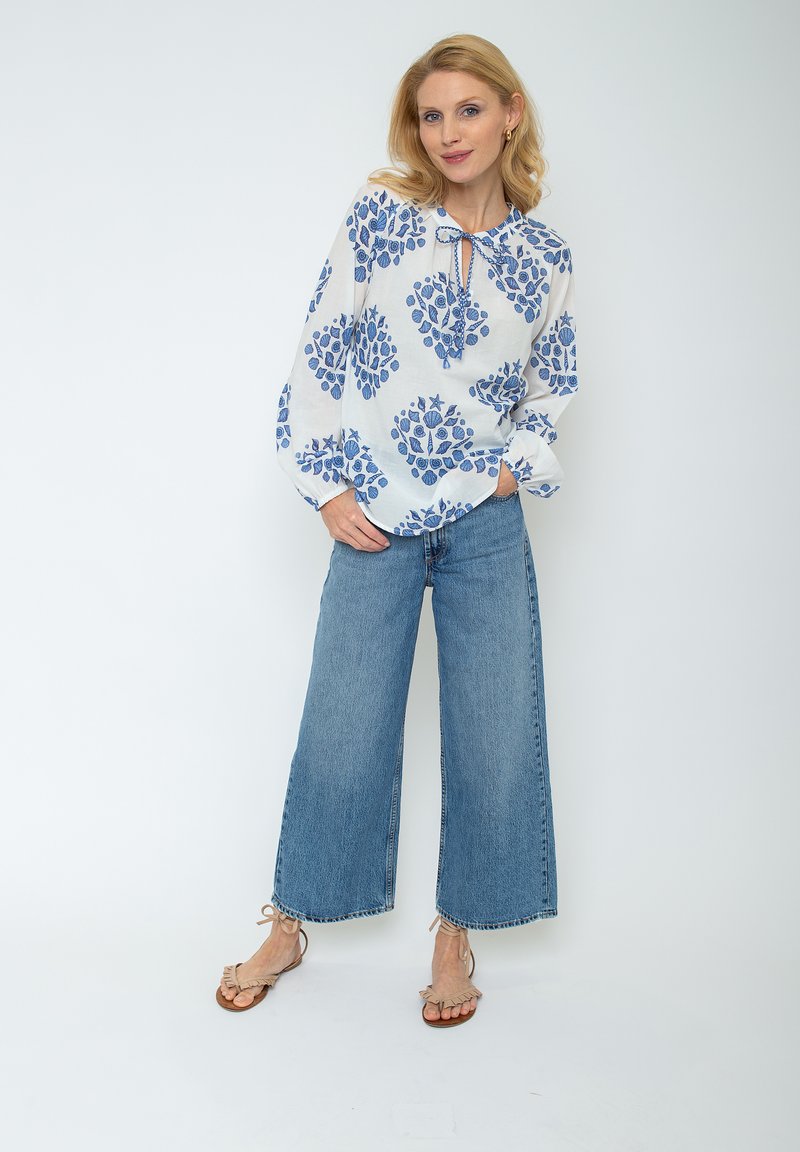 Biała bluzka w niebieski kwiatowy wzór, luźny krój, z wiązanym dekoltem; zestawiona z niebieskimi spodniami o szerokich nogawkach i beżowymi sandałami z falbanami.