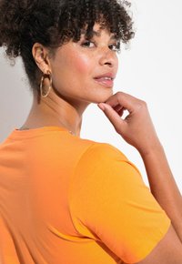 Kortærmet orange T-shirt lavet af blødt stof, med en rund halsudskæring og en glat tekstur, med en metallisk detalje nær skulderen.