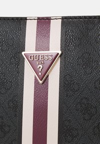 Juodas tekstūruotas krepšys su bordo ir kreminėmis vertikaliomis juostelėmis. Yra auksinė trikampė logotipo etiketė su "GUESS USA" tekstu ir klausimo ženklu.