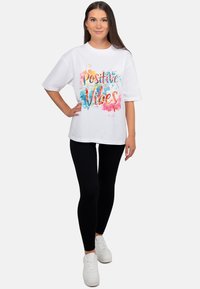 Wit katoenen t-shirt met de tekst "Positive Vibes" in een kleurrijke, aquarelachtige print, gecombineerd met zwarte legging en witte sneakers.