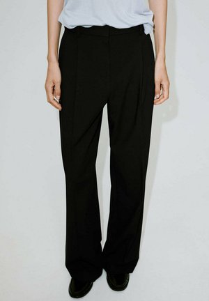 Persona che indossa pantaloni neri larghi e scarpe nere, in piedi davanti a uno sfondo chiaro uniforme, mani rilassate lungo i fianchi.