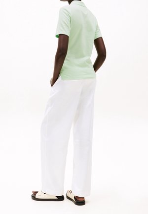 Personne portant une chemise à manches courtes vert clair, un pantalon blanc ample et des sandales beiges, debout les mains dans les poches sur fond blanc.