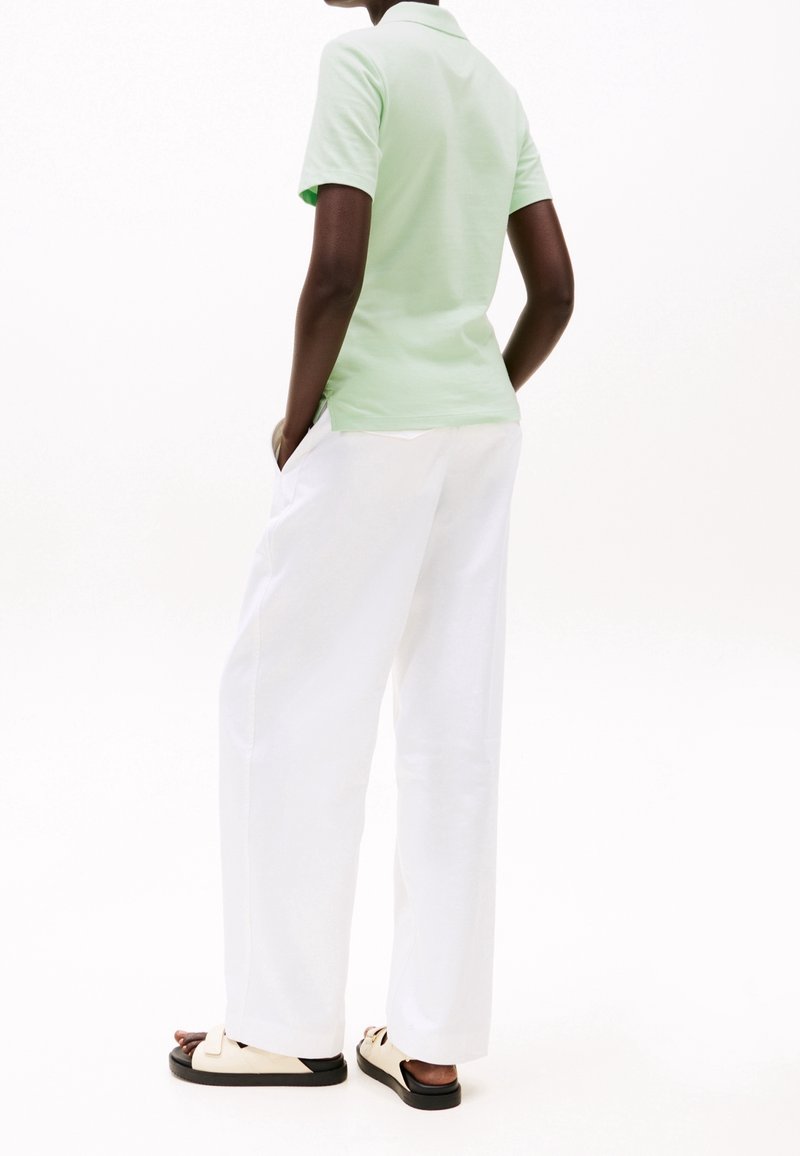 Personne portant une chemise à manches courtes vert clair, un pantalon blanc ample et des sandales beiges, debout les mains dans les poches sur fond blanc.