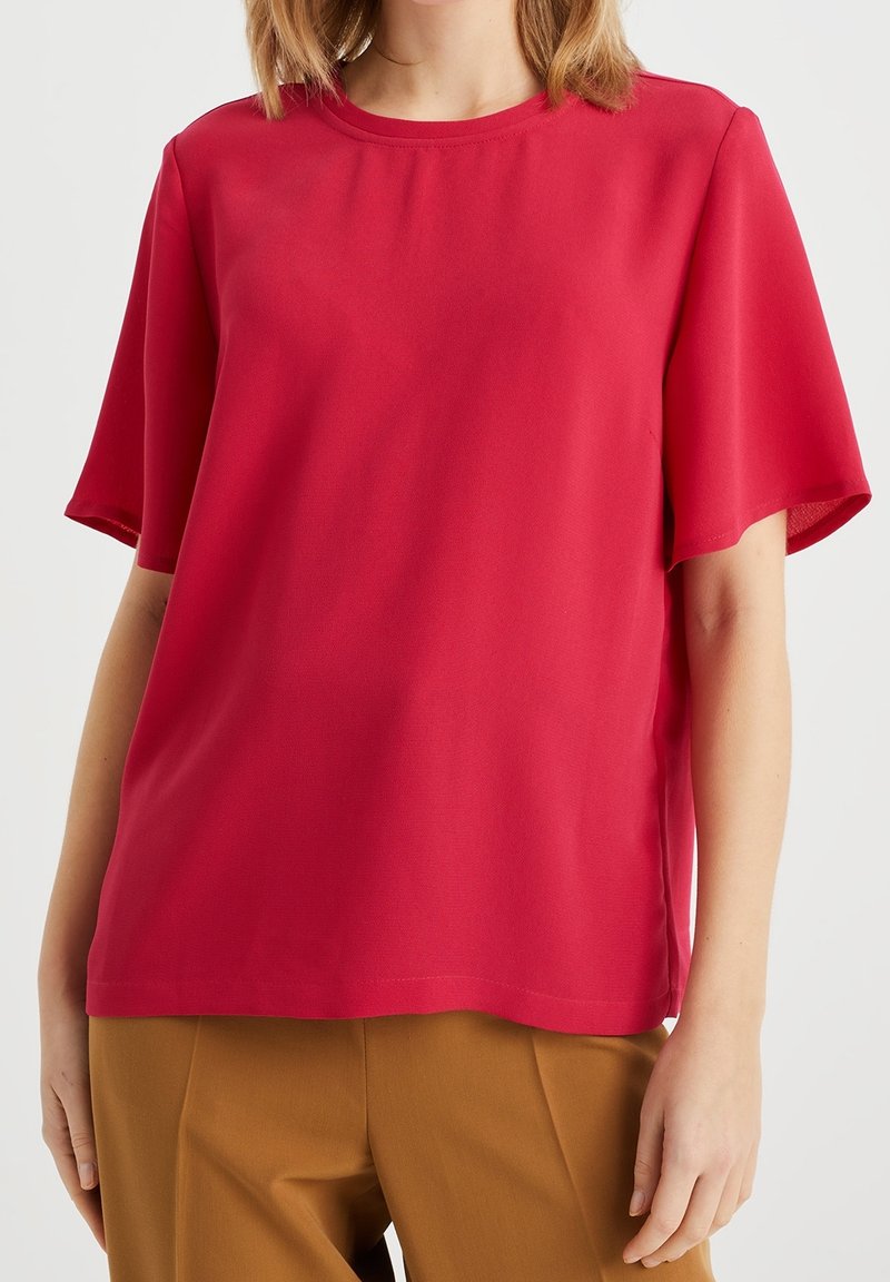 Top rosso a maniche corte realizzato in tessuto morbido con scollatura rotonda. Vestibilità ampia e design semplice, senza motivi o decorazioni visibili.