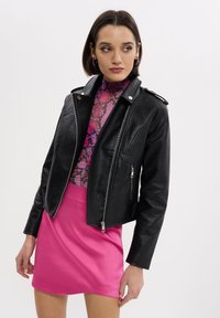 Donna che indossa una giacca di pelle nera, un top rosa con motivo a serpente e una gonna rosa brillante, che guarda verso sinistra su uno sfondo neutro.