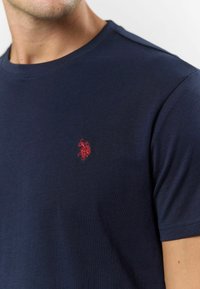 Námořnická modrá bavlněná tričko s krátkými rukávy, s malým vyšívaným červeným logem na hrudi. Hladká textura, kulatý výstřih.