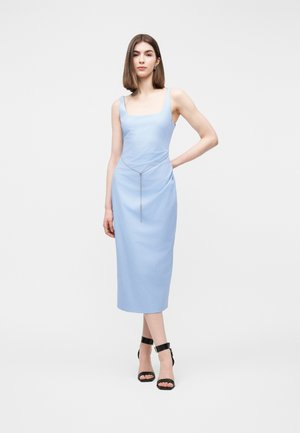 Junge Frau steht mit der Hand in der Hüfte, trägt ein ärmelloses, hellblaues Midikleid, einen Kettengürtel und schwarze Schuhe mit Knöchelriemen vor weißem Hintergrund.