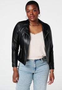 Veste de biker en simili cuir noir avec fermeture éclair à l'avant, détails matelassés sur les épaules, associée avec un haut beige à côtes et un jean bleu clair.