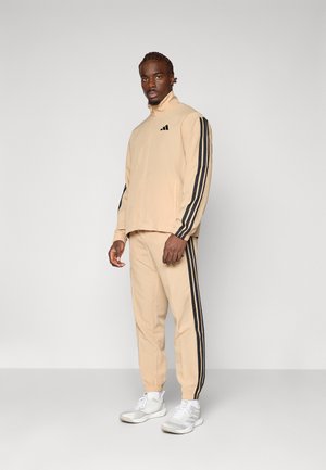 Beige tracksuit with black side stripes; zip jacket with high collar, elastic cuffs; matching pants with elastic ankles; paired with white sneakers.