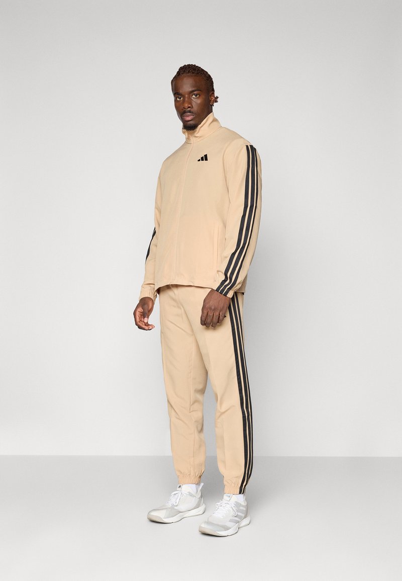 Survêtement beige avec des bandes latérales noires ; veste zippée à col montant, poignets élastiques ; pantalon assorti avec chevilles élastiques ; associé à des baskets blanches.