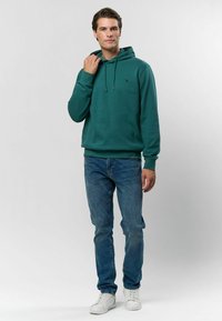 Zöld kapucnis pulóver kenguru zsebbel, húzózsinórral és logóval. Kék farmernadrággal és fehér sportcipővel párosítva. Sima textúra.