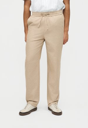 Férfi bézs színű, húzózsinóros nadrágban és fehér tornacipőben, fehér háttér előtt áll, kezei nyugodtan oldalra eresztve.