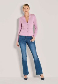 Frau in einer helllila, knöpfbaren Strickjacke, blauen Bootcut-Jeans und dunklen spitzen Absätzen, die vor einer schlichten hellen Wand steht.