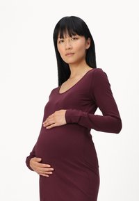 Sötét bordó színű kismama ruha, testhez álló fazonnal, hosszú ujjakkal, puha anyagból készült, és mély dekoltázsú, finoman öleli körül a sziluettet.