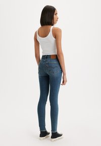 Jeans ajustados en denim azul oscuro, con una cintura alta, bolsillos traseros y un parche de cuero marrón en la cinturilla, combinados con una camiseta sin mangas blanca.