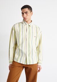 Camisa de botones a rayas que presenta colores amarillo, verde y blanco, con dos bolsillos en el pecho y un parche con el logo. Combinada con pantalones marrones.