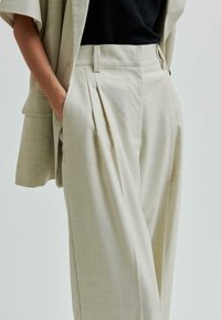Pantaloni plissettati color beige realizzati in tessuto leggero, con vita alta, tasche laterali e un fit rilassato. Texture liscia con dettagli sartoriali.