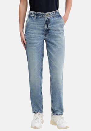 Persona che indossa jeans a vita alta, a gamba dritta di colore azzurro chiaro, sneakers bianche, con una mano in tasca, su sfondo semplice.